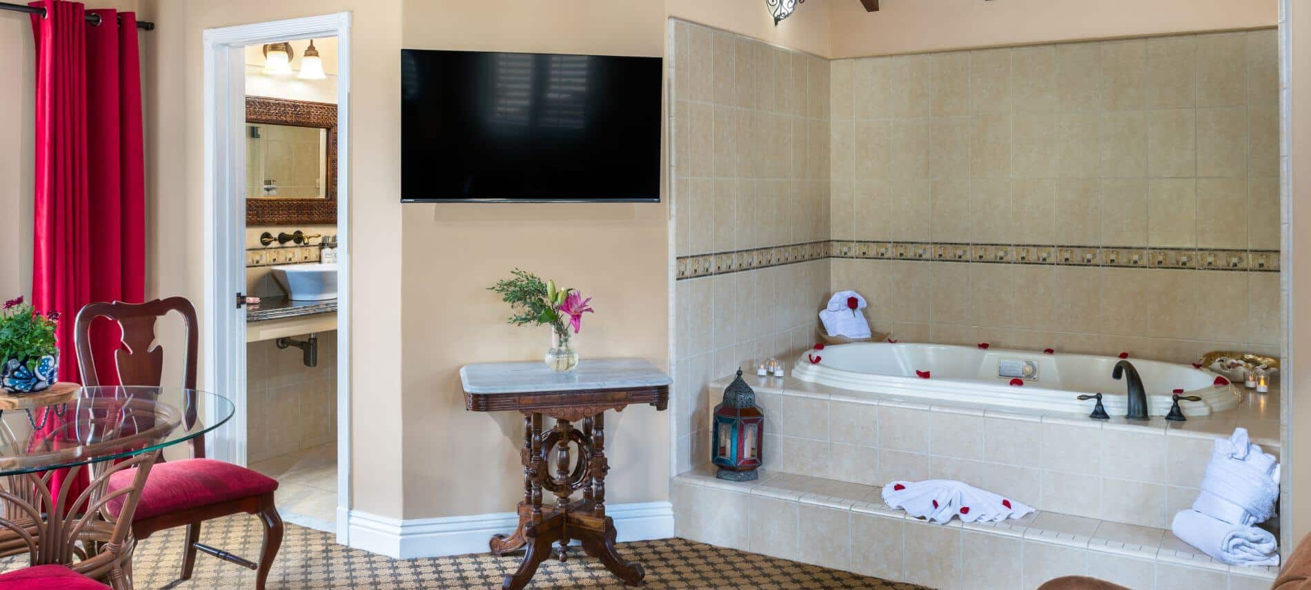bathtub in a tiled cubby hole, flat tv on wall, glass table with chairs, red curtains