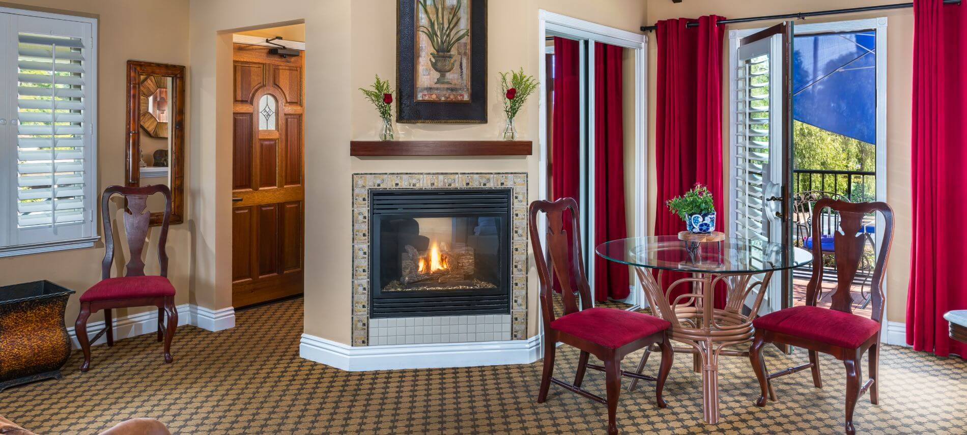fireplace in middle of room inset in the wall, glass table with wood red velvet cushioned chairs, open door to patio