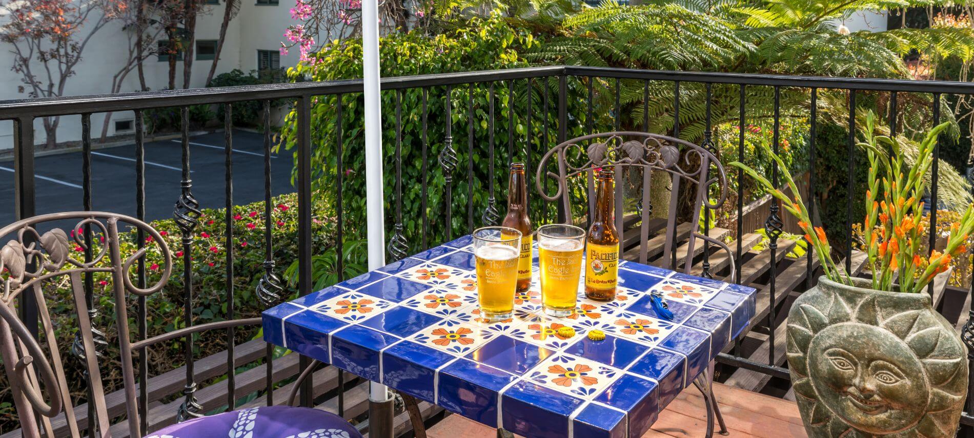 patio surrounded by foliage with blue and white checkered table, blue cushioned wrought iron chairs, beer glasses and bottles