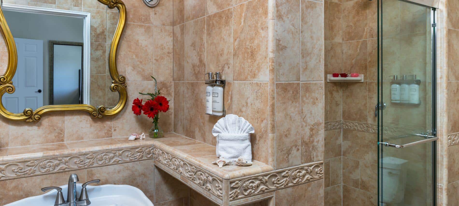 L shaped counter tiled shelf above white sink under gold trimmed mirror on wall, walk in shower with glass door