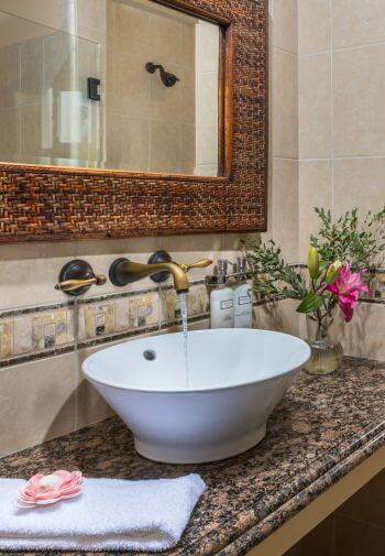 white sink on top of marble shelf with wicker framed mirror with reflection of shower, flowers in corner, white towel on shelf