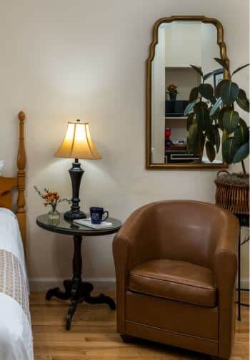 brown leather horseshoe shaped chair under gold framed mirror beside bed, coffee mug on stand with lamp