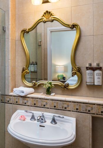 cream tiled bathroom,white pedestal sink with gold trimmed mirror hanging above with reflection of bedroom in glass