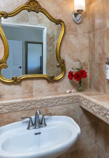 tiled room with white sink with chrome fixtures under gold trimmed mirror with light on wall to right