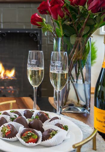 two champagne glasses with the eagle inn logo behind a plate of chocolate covered strawberries with red roses in vase behind