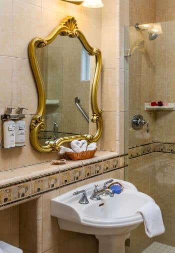 white pedestal sink under gold framed mirror, glass enclosed tiled shower with grab bar