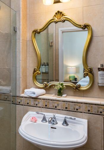 cream tiled bathroom,white pedestal sink with gold trimmed mirror hanging above with reflection of bedroom in glass